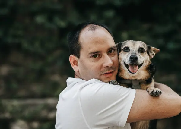 Beyaz tişörtlü bir adam küçük kahverengi ve siyah bir köpek tutuyor. Arka plan bulanık ve bitki örtüsü olan bir açık hava ortamında.