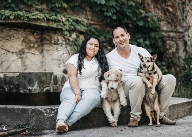 Taş duvarda iki köpekle oturan bir çift. İkisi de beyaz elbiseler ve kot pantolon giyiyor. Soldaki köpek bir tilki teriyeri, sağdaki köpeğin ise kısa koyu bir paltosu var..