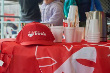 Üzerinde Teleton logosu olan kırmızı masa örtüsü olan bir masa. Üzerinde kep, bardak, çaydanlık ve şeker torbaları var. Arka planda, insanlar bir hayır kurumunda ve halk etkinliğinde dikkatlerini dağıtıyor..