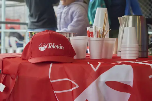 Üzerinde Teleton logosu olan kırmızı masa örtüsü olan bir masa. Üzerinde kep, bardak, çaydanlık ve şeker torbaları var. Arka planda, insanlar bir hayır kurumunda ve halk etkinliğinde dikkatlerini dağıtıyor..