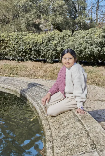 Parktaki taş çeşmenin yanında pembe beyaz bir kazak giyen Latin bir kızın portresi. Doğal ışık ve sakin ifade çocukluk, kimlik ve çeşitlilik kavramları için idealdir..