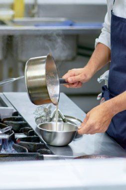 Chef pours hot liquid into metal bowl. Visible steam, active professional kitchen, precise culinary technique, and authentic gastronomic atmosphere.