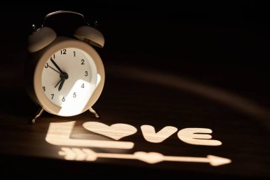 Retro tarzı alarm saati, beyaz ışıkla aydınlatılmış, komodinin üzerinde Aşk ve Aşk Tanrısı 'nın oku yazıyor.