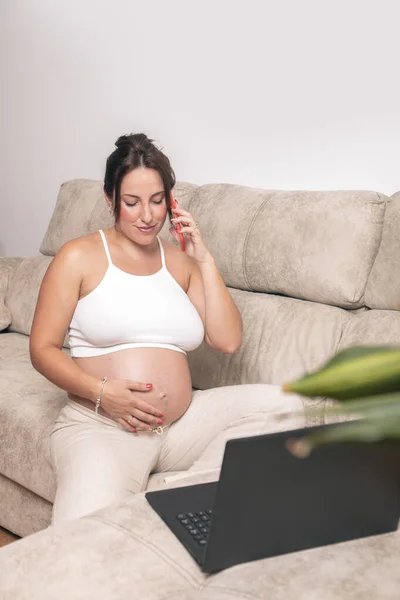 Genç Hamile Bir Kadın Telefondayken Karnına Dokunurken Evdeki Kanepesinde Dizüstü — Stok fotoğraf