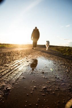 Bir adam ve köpeği toprak bir yolda yürüyorlar. Adam ceket giyiyor ve köpek tasma takıyor.