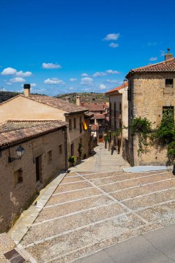 Sakin, dar bir cadde taştan binalar ve renkli bayraklarla kaplı küçük bir İspanyol köyünden geçiyor. Mavi gökyüzü konumun huzurlu atmosferine eklenir..