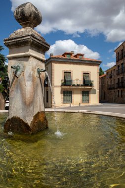 Ön plandaki tarihi bir fıskiyeden nazikçe akan su, açık bir günde klasik mimarisi olan sakin bir köyün atmosferini vurguluyor..