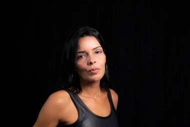Studio portrait of young woman looking at camera against simple studio black background. She wears a black t-shirt.