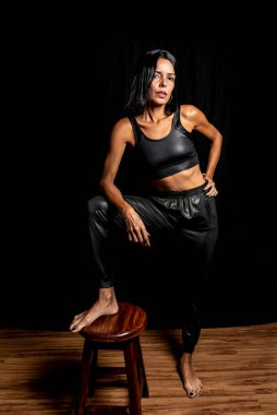 Siyah tişörtlü ve pantolonlu genç bir kadının stüdyo portresi. Ayağı tahta bir bankta, siyah bir stüdyo arka planında. Salvador, Brezilya.
