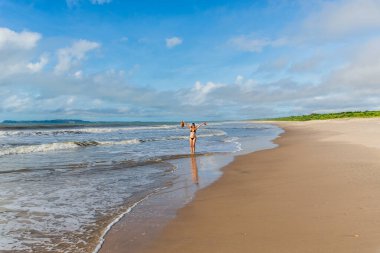 An adult woman in a bikini walking under strong sunlight on Guaibim beach in the city of Valenca, Brazilian state of Bahia. Travel and fun.