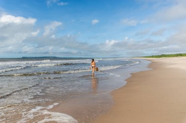 An adult woman in a bikini walking under strong sunlight on Guaibim beach in the city of Valenca, Brazilian state of Bahia. Travel and fun.