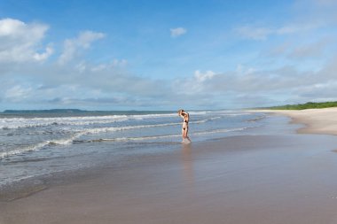 An adult woman in a bikini walking under strong sunlight on Guaibim beach in the city of Valenca, Brazilian state of Bahia. Travel and fun.