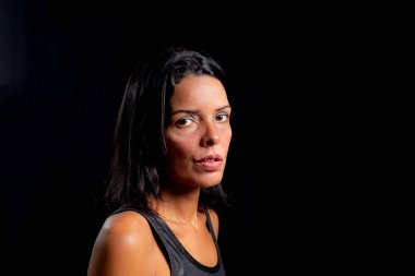 Studio portrait of young woman looking at camera against simple studio black background. She wears a black t-shirt.