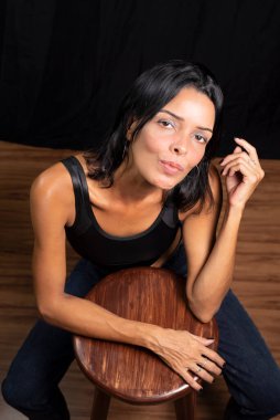 Siyah tişörtlü genç bir kadının siyah bir stüdyo arka planı ve ahşap zemine bakan bir stüdyo portresi. Salvador, Brezilya.