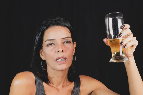 Portrait of young woman in black t-shirt drinking beer from glass cup. against black studio background. Salvador, Brazil.