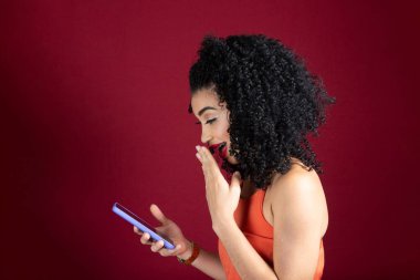 Genç, güzel, neşeli bir kadın cep telefonuna bakıyor. Turuncu giysi giyiyor. Kırmızı stüdyo arka planında izole edilmiş..
