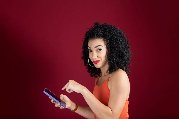 stock image Young beautiful cheerful woman pointing at cellphone and looking at camera. wearing orange clothing. Isolated on red studio background.