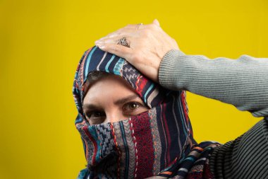 Başörtüsü takan kadın portresi, kameraya bakıyor, elleri başının üstünde, sarı stüdyo arka planında izole edilmiş..