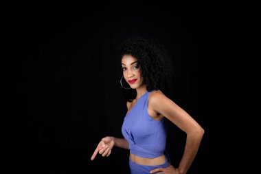 Genç, güzel, gülümseyen bir kadın parmağıyla işaret ediyor. Mavi elbiseler giyiyordu. Stüdyoda siyah arkaplan.