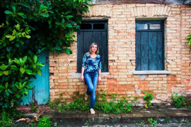 Eski bir evin camına yaslanmış bir kadın. Tuğla cephesi ve etrafta dolaşır. Aratuipe, Bahia.