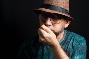Şapkalı ve gözlüklü bir adam, eli ağzında. Koyu arkaplanda izole.