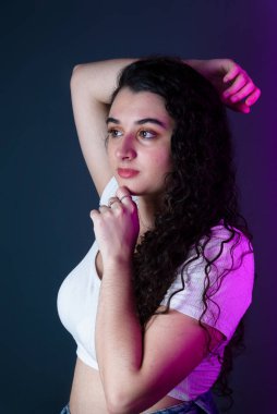 Bir eli başında, diğer eli çenesinde olan beyaz tişörtlü genç bir kadın profili. Gri arkaplanda izole.