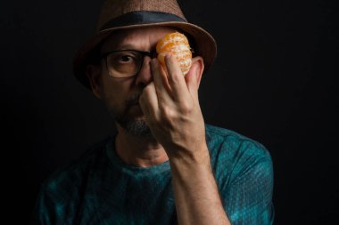 Elinde mandalina olan şapkalı ve gözlüklü bir adam. Koyu arkaplanda izole.