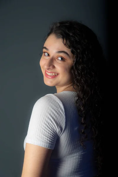 Profile of beautiful young woman with black hair wearing white shirt and looking at camera. Against gray background.