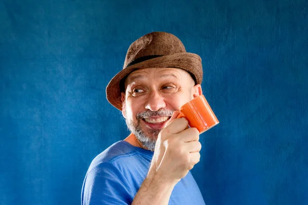 Funny smiling man wearing a hat holding an orange cup. Isolated on blue background