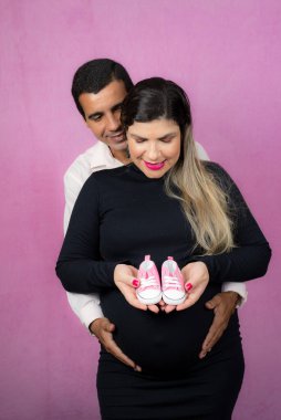 Romantic couple of handsome husband and beautiful pregnant wife posing in studio. She holds baby slippers.