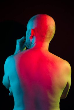 Portrait of bald man, shirtless and back to the camera, with colorful lights of studio. Isolated in the black background.