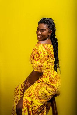 Beautiful young woman sitting on a stool. Colorful clothes. Isolated on yellow background.