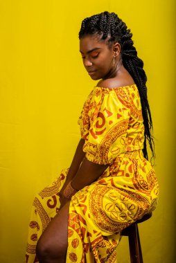 Beautiful young woman sitting on a stool. Colorful clothes. Isolated on yellow background.