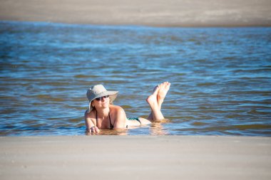 Bikinili ve şapkalı bir kadın denizin sularında ayaklarını kaldırıyor. Taquari, Guaibim plajı, Valenca, Bahia.