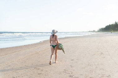 Bikinili ve şapkalı güzel bir kadın, arkadan kumsalda yürüyor. Guaibim Plajı, Valenca, Bahia.