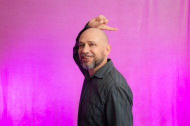 Ciddi bir adamın portresi, 50 yaşında, sakallı ve kel kafalı, el işaretleri yapan. Pembe arka planda izole edilmiş.