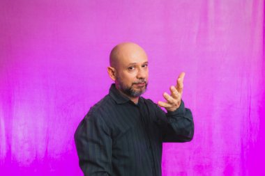 Ciddi bir adamın portresi, 50 yaşında, sakallı ve kel kafalı, el işaretleri yapan. Pembe arka planda izole edilmiş.