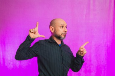 Ciddi bir adamın portresi, 50 yaşında, sakallı ve kel kafalı, el işaretleri yapan. Pembe arka planda izole edilmiş.