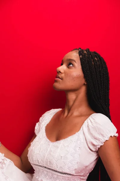 stock image Beautiful young black woman sitting and serene wearing white outfit with long braids in her hair. Isolated on red background