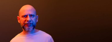 Sakallı kel bir adam, ciddi, kameraya şüpheli bir şekilde bakıyor. Düşünceli ve odaklanmış. Koyu kırmızı arkaplanda izole.