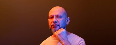 Sakallı kel bir adam, ciddi, kameraya şüpheli bir şekilde bakıyor. Düşünceli ve odaklanmış. Koyu kırmızı arkaplanda izole.