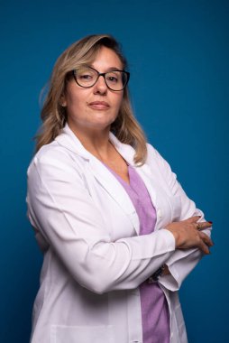 Beyaz ceket ve leylak bluz giymiş, kameraya bakan tatlı, kendinden emin orta yaşlı bir hemşirenin fotoğrafı. Mavi arkaplanda izole edilmiş