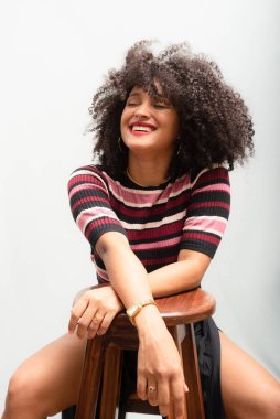 Tahta bir taburede oturan güzel, gülümseyen, kıvırcık saçlı genç bir kadının portresi. Açık gri arkaplanda izole.