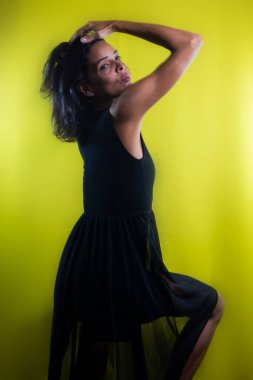 Uzun siyah elbiseli güzel bir manken, ahşap bir taburede oturuyor, eli başının üstünde ve kameraya bakıyor. Sarı arkaplanda izole edilmiş.