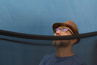 Siyah şeffaf bir kumaşın arkasında şapka, gözlük ve sakal takan bir adam. Mavi arkaplanda izole edilmiş.