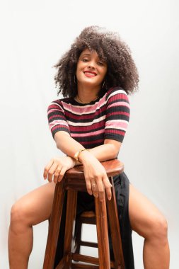 Tahta bir taburede oturan güzel, gülümseyen, kıvırcık saçlı genç bir kadının portresi. Açık gri arkaplanda izole.