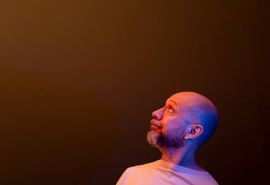 Sakallı kel bir adam, ciddi, şaşırmış bir ifadeyle yukarı bakıyor. Koyu kırmızı arkaplanda izole.