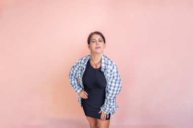 Çekici esmer, siyah elbiseli, kareli bluzlu, ayakta kameraya poz veriyor. Stüdyo portresi. Somon arka planında izole edilmiş.