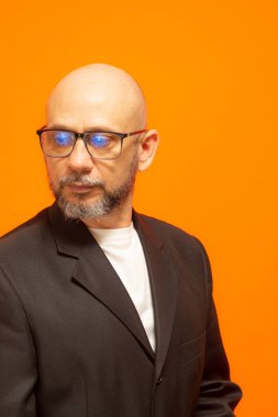 Takım elbiseli, sakallı, kel ve gözlüklü şık bir adam. Turuncu arka planda izole edilmiş