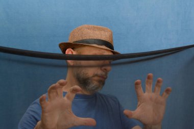 Şeffaf siyah kumaşın arkasında şapkalı ve sakallı bir adam var. Mavi arkaplanda izole.